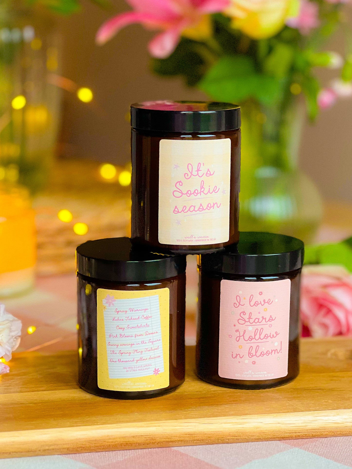 Three candles with labels on a wooden surface with flowers in the background